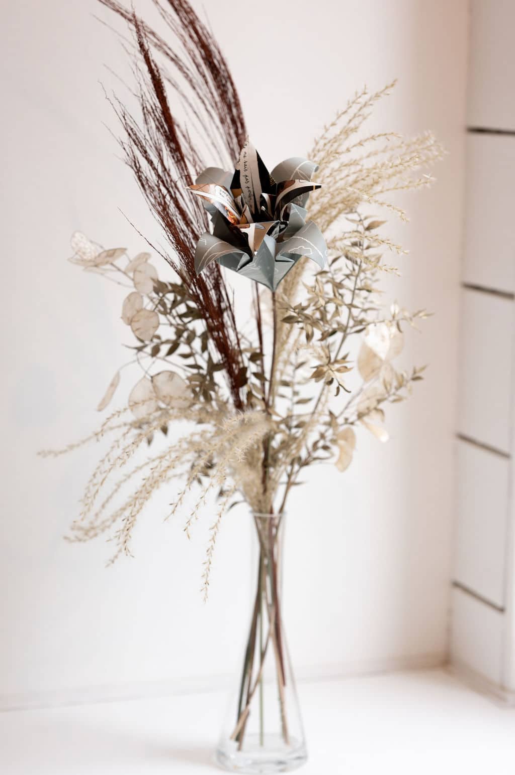Cette photo représente une grande et haute composition florale comprenant une fleur de papier en forme de double lys et des fleurs séchées dans les tons marrons et beige