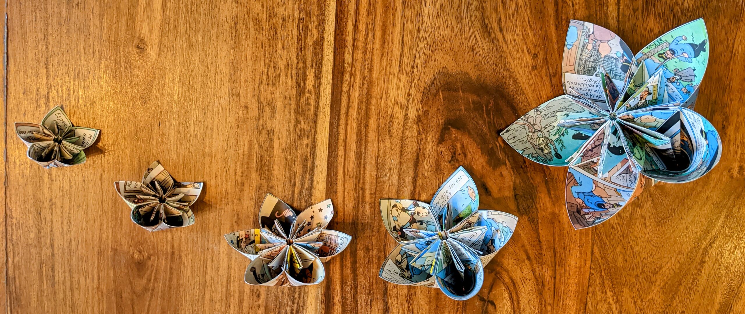 Cette photo représente une ribambelle de fleurs de papier sans tige posées en arc de cercle sur une table en bois. Ces fleurs de papiers sont réalisées à partir d'une BD de tintin