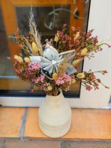 Élégant bouquet mixte composé de fleurs séchées aux tons pastel et d'une fleur en papier pliée à partir de plusieurs pages de livre ancien, le tout arrangé dans un vase en céramique écru.