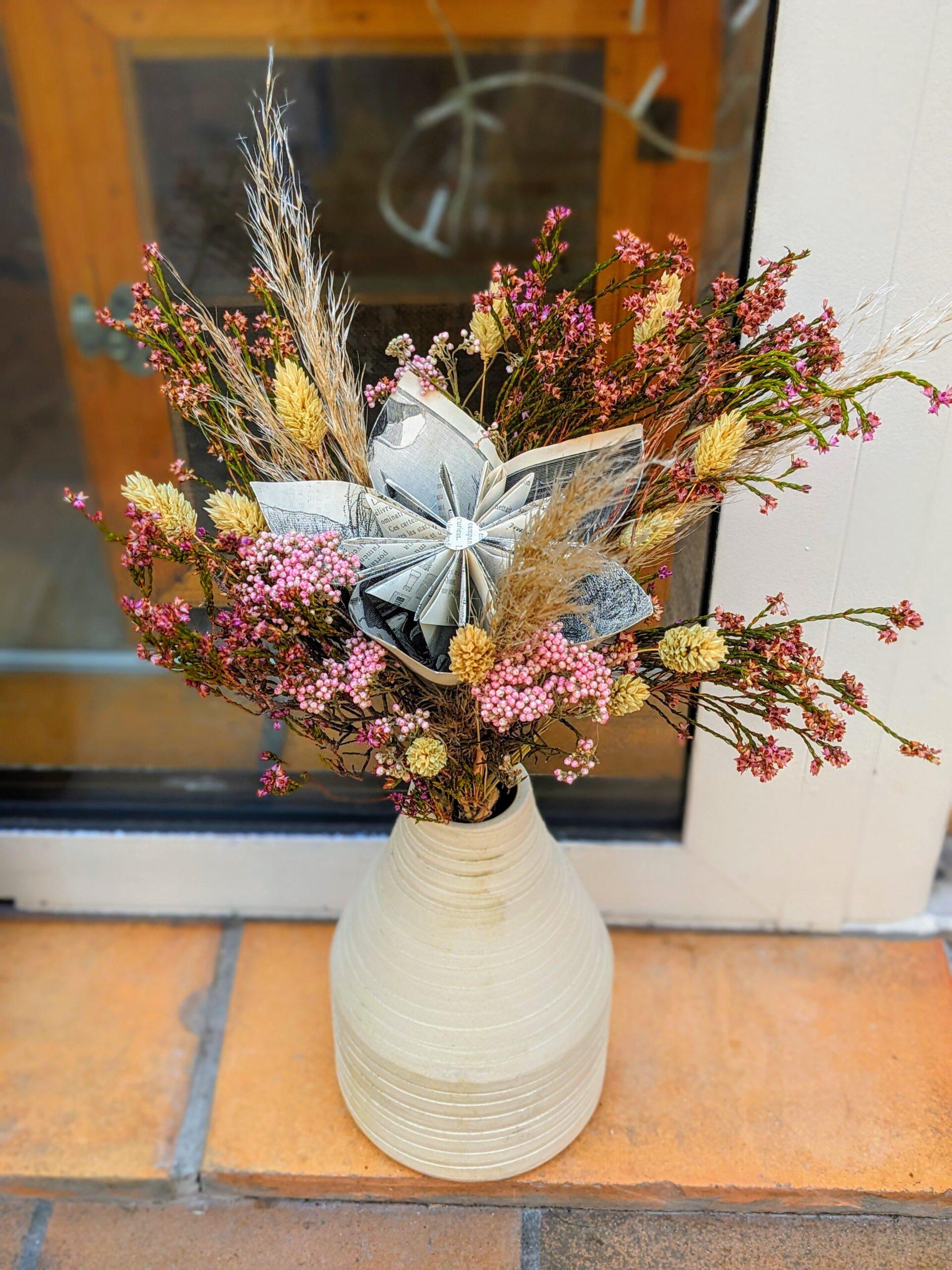 Élégant bouquet mixte composé de fleurs séchées aux tons pastel et d'une fleur en papier pliée à partir de plusieurs pages de livre ancien, le tout arrangé dans un vase en céramique écru.